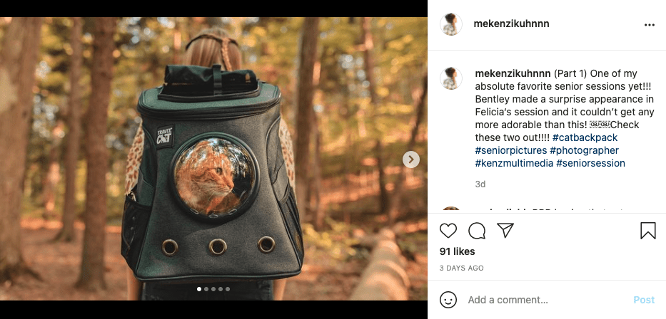 woman wearing a black Travel Cat backpack. In the backpack is an orange cat. The background is a forest.