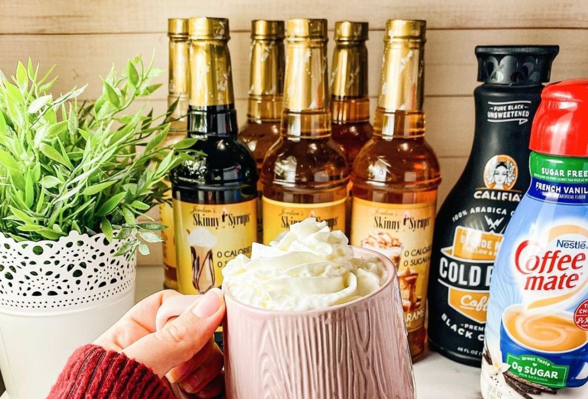 Woman holding mug in front of a bunch of bottles of Skinny Mixes Coffee Syrup